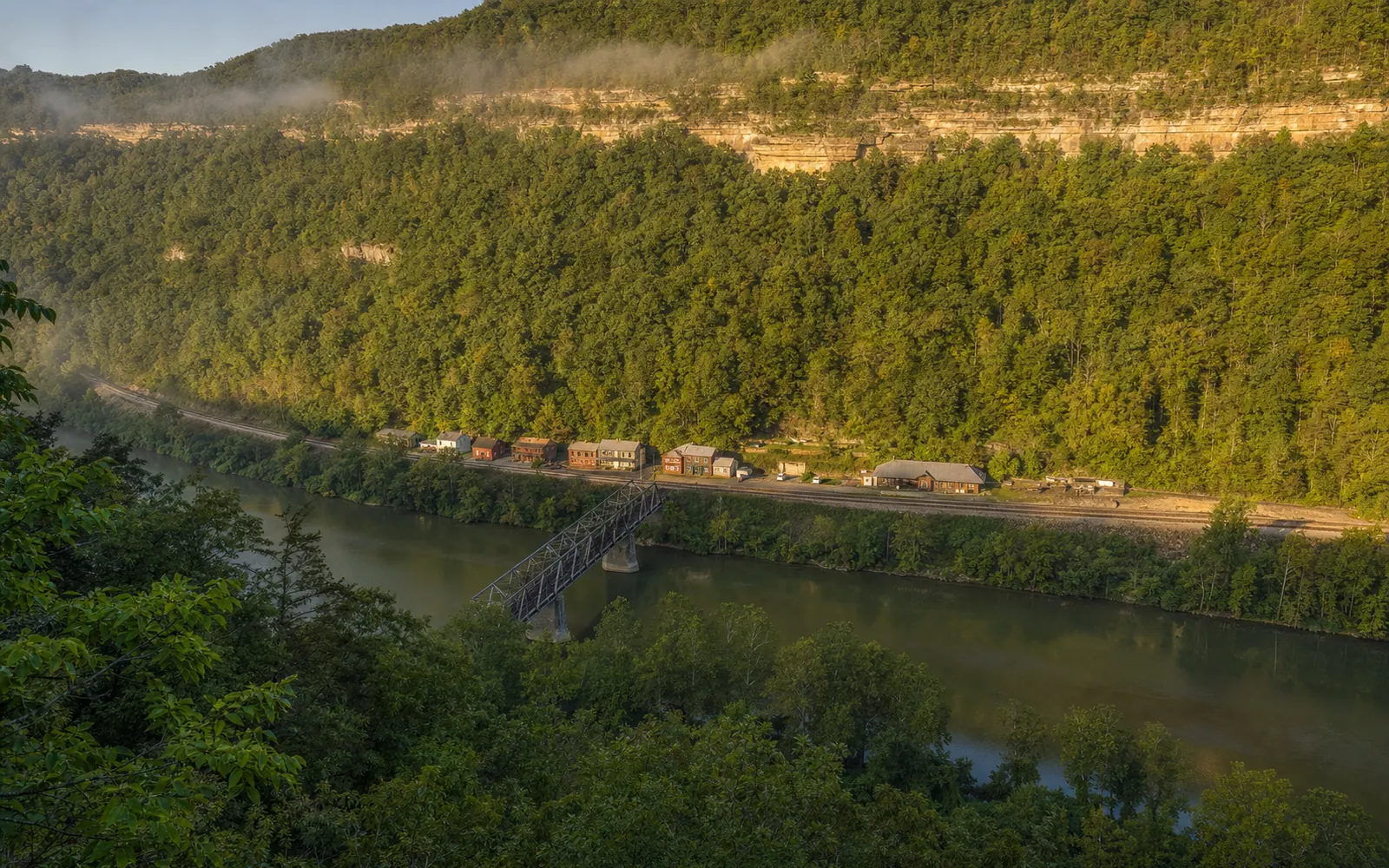 Thurmond seen from the hillside above, a short row of buildings and the depot on a narrow shelf between the railroad tracks and the river, a truss bridge crossing to the town, AI-generated illustration