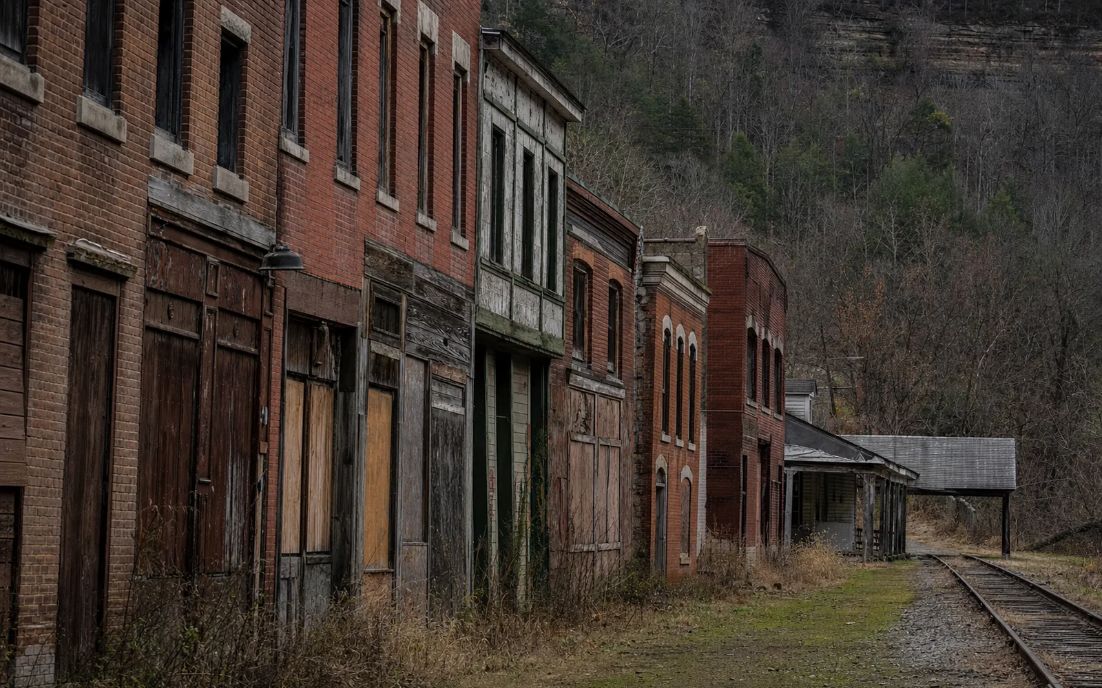 Thurmond's weathered brick and wood commercial row with boarded doors and dry weeds beside the railroad tracks, the depot's long roof at the far end under a wooded cliff, AI-generated illustration