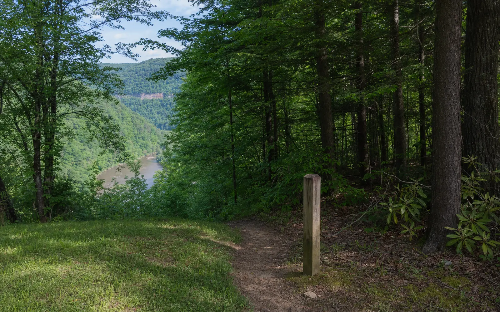 A narrow footpath leaves the mown edge of a town park into hemlock woods past a wooden trail post, with the ground falling away toward the gorge, AI-generated illustration