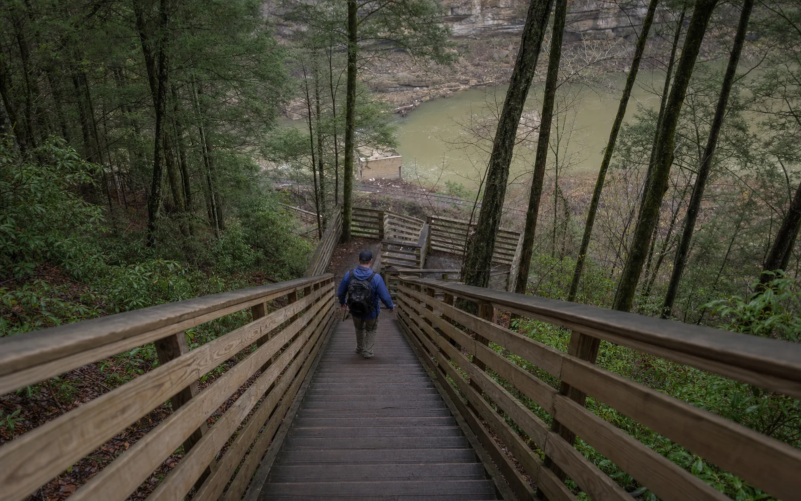The wooden stairs of the Kaymoor Miners Trail descend steeply through hemlocks, with an adult hiker on the steps and mine ruins below, AI-generated illustration
